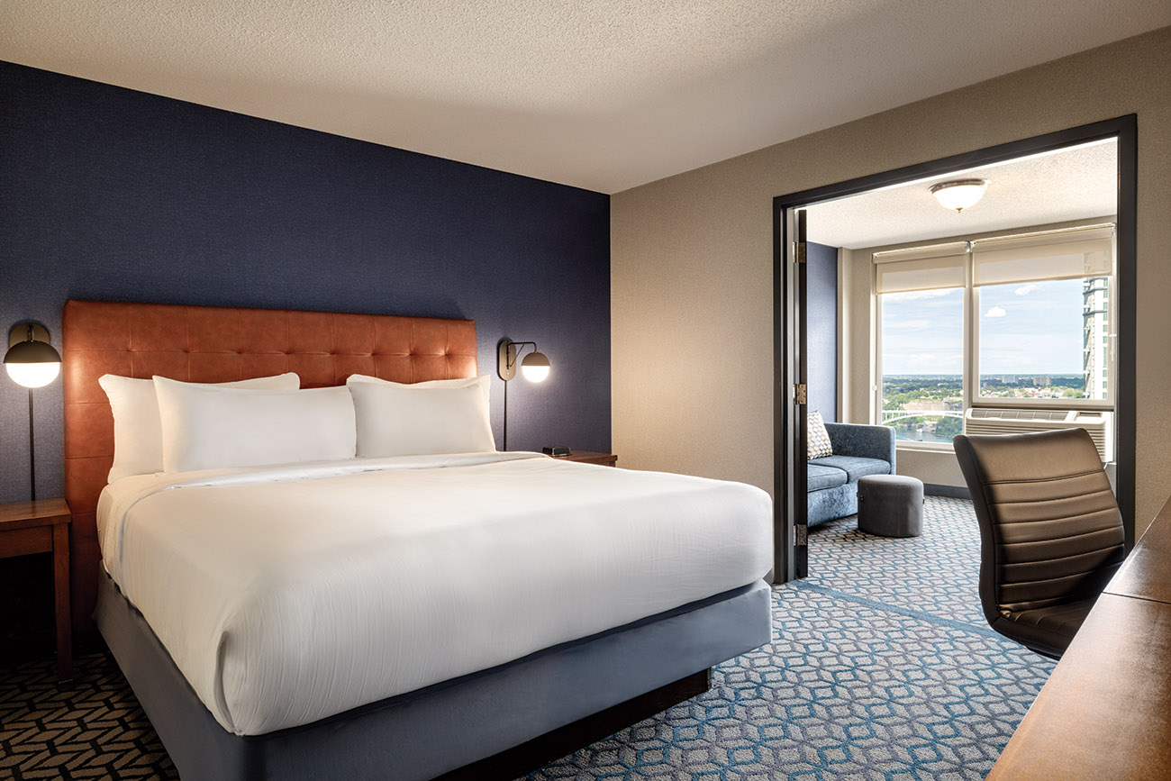 A hotel room with a king bed with white linen, open to a second room with a seating area with couch and large window.